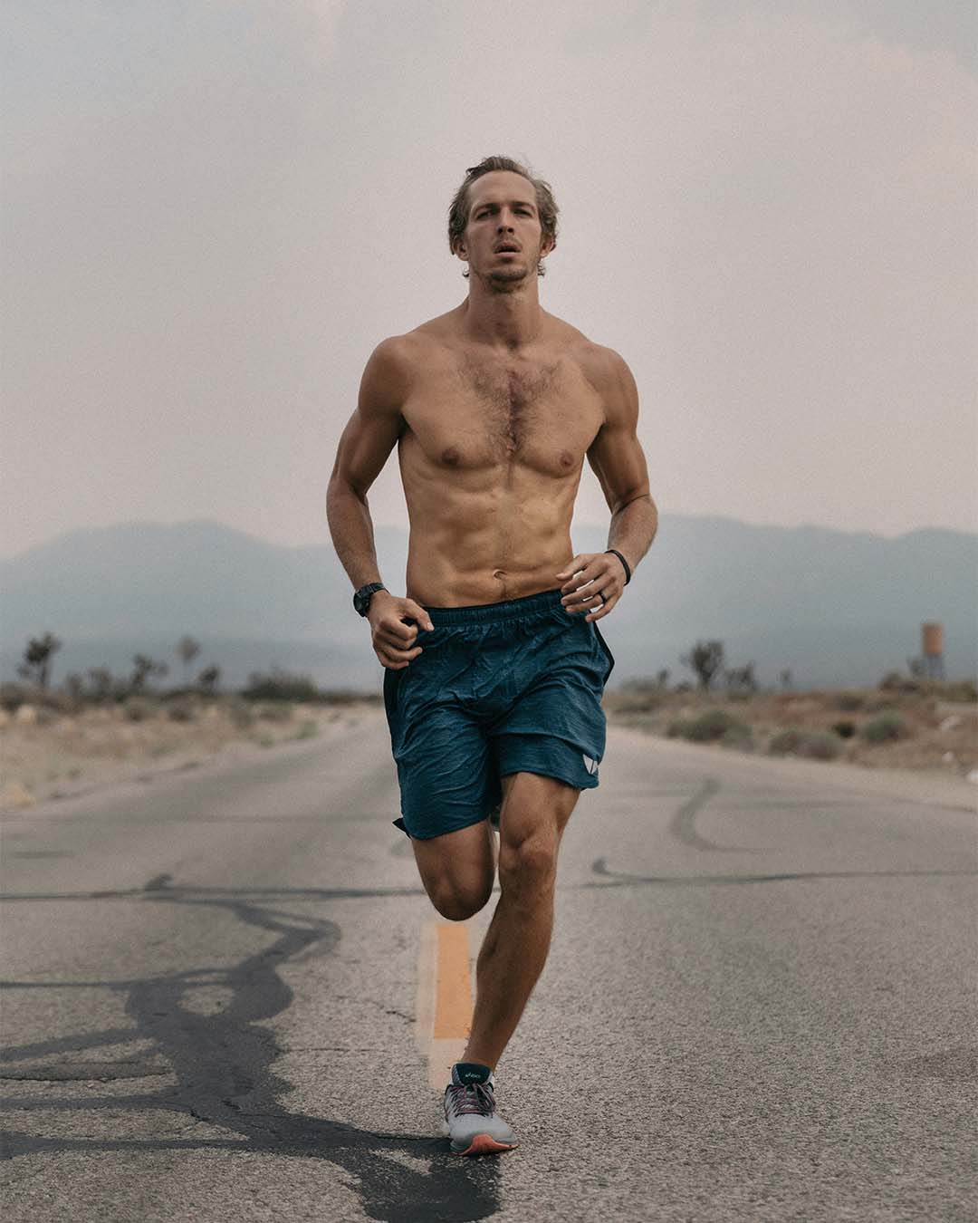 Deportista sin camisa, de complexión musculosa, corriendo por una carretera desierta con montañas al fondo.