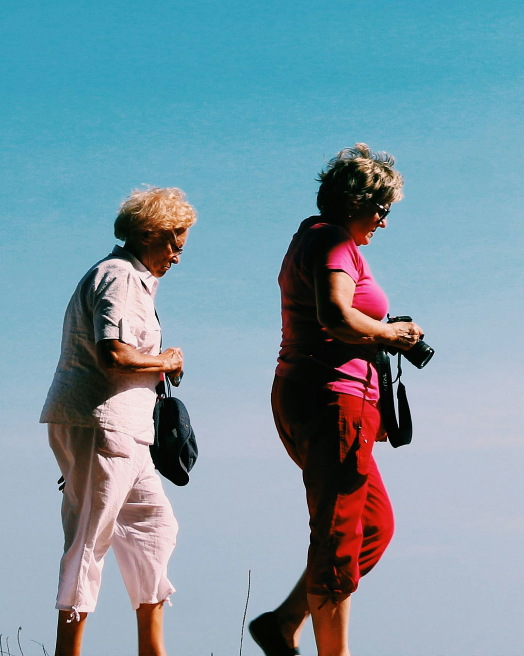 Dos mujeres de la tercera edad caminando al aire libre bajo un cielo azul claro, una sosteniendo una cámara y la otra llevando un sombrero.