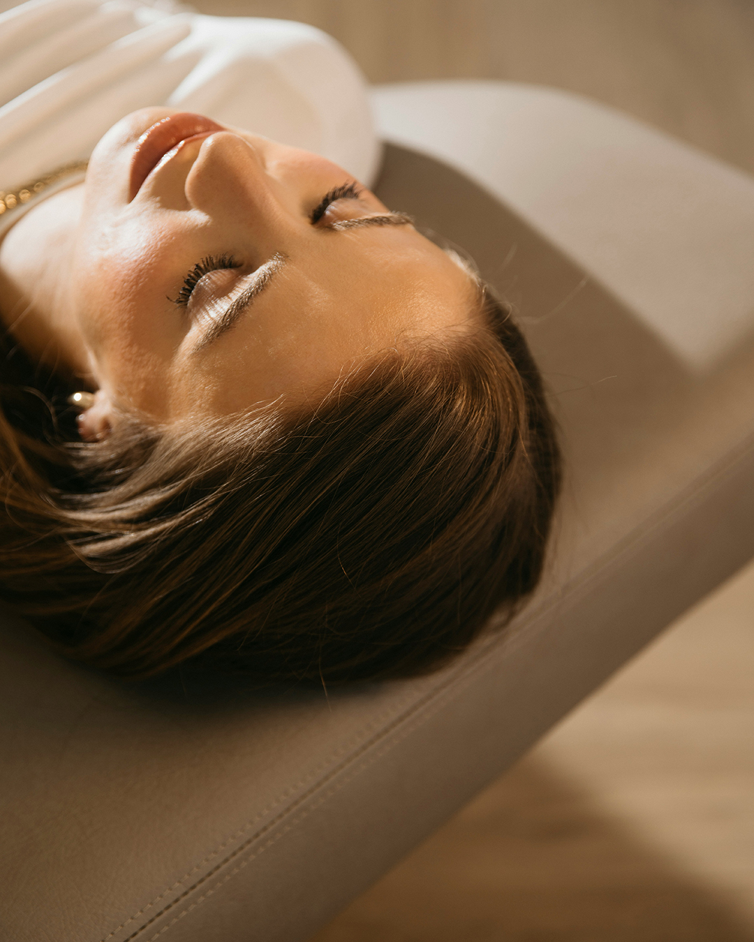 Primer plano de una mujer con los ojos cerrados, tumbada sobre una superficie acolchada de color gris, con aspecto relajado después de recibir un tratamiento de recuperación.